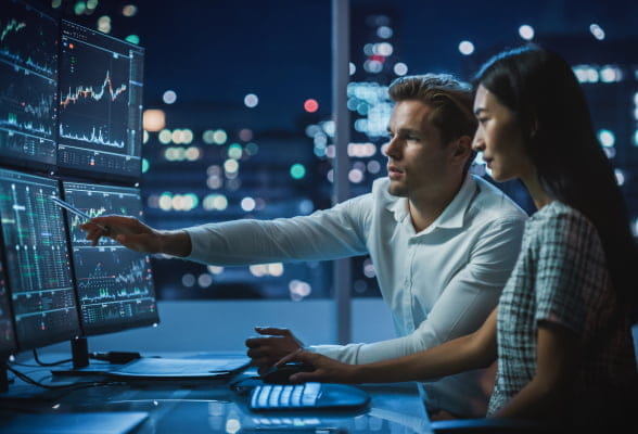 Two users looking, while one is pointing,  at a multi-monitor trading setup for StoneX PRO solutions